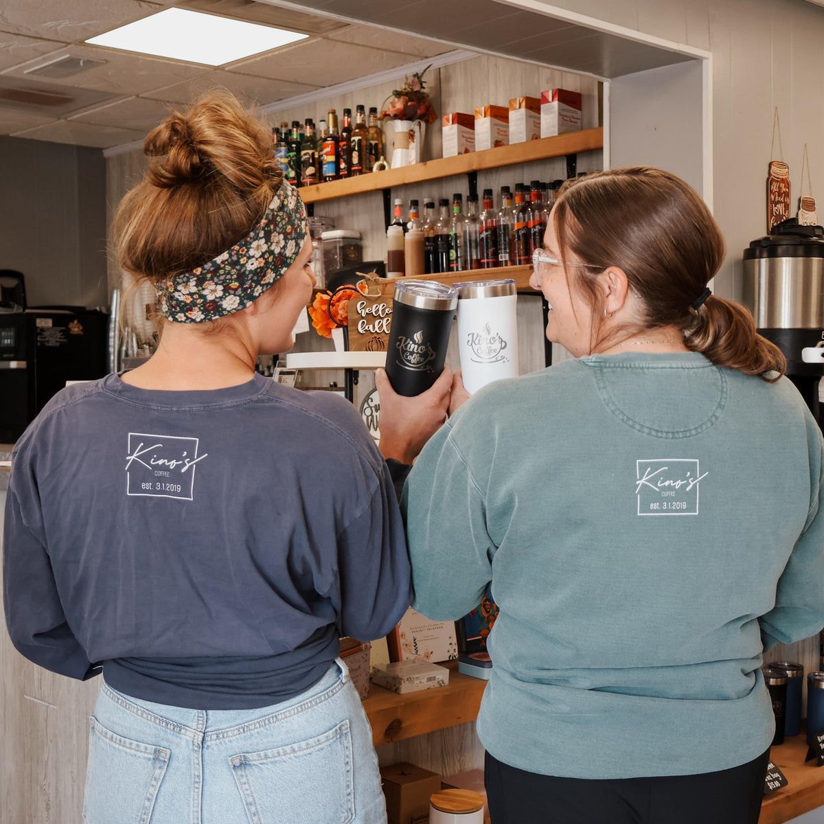 Two women in a kitchen or bar setting, one holding a black cup.
